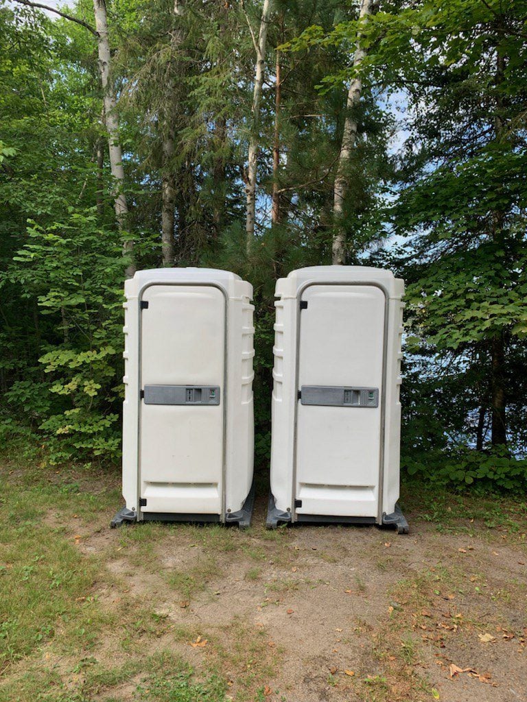 Two portable toilets in a wooded area by a lake
