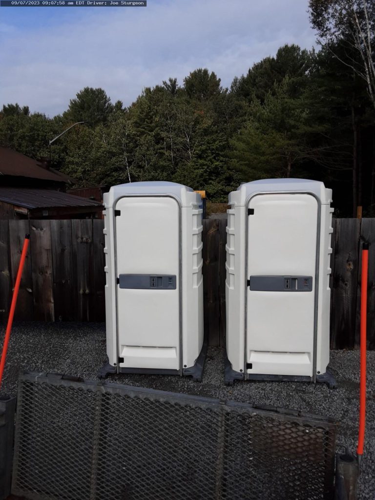 Two portable toilets on gravel with wooden fence backdrop